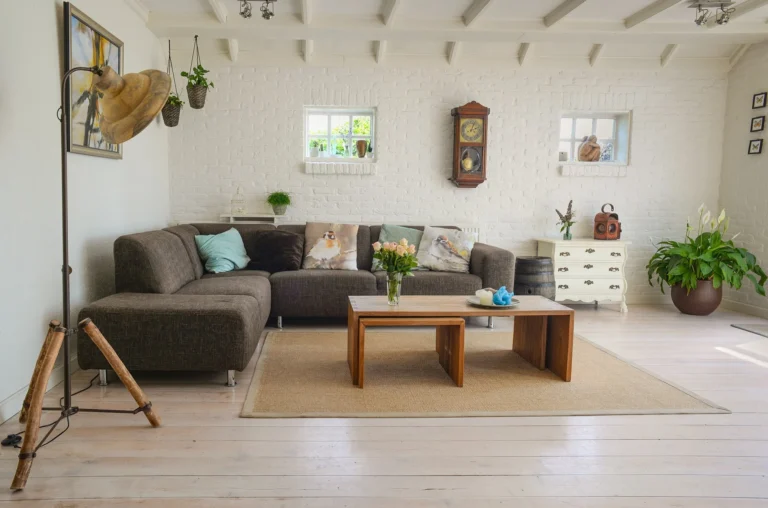 A bright and cozy living room with a grey L-shaped sofa, nested wooden coffee tables on a jute rug, whitewashed brick walls, a vintage wall clock, and houseplants.